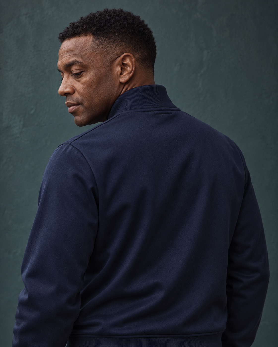 Rear close-up of a man wearing the navy Track Jacket Heritage Signature, looking over his shoulder against a teal wall